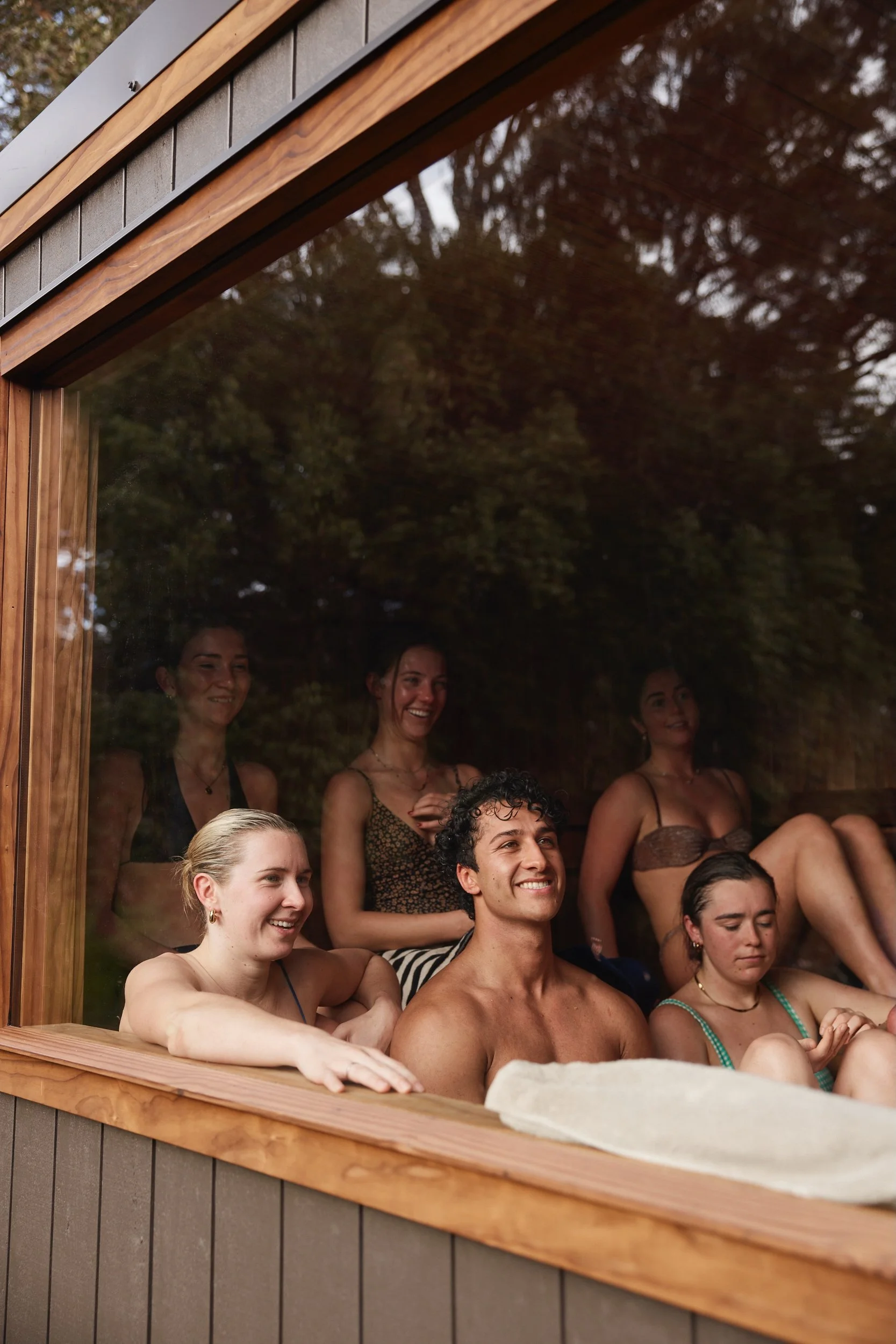 People relaxing and smiling inside a wooden structure with a glass window, outdoor trees reflected in the glass.