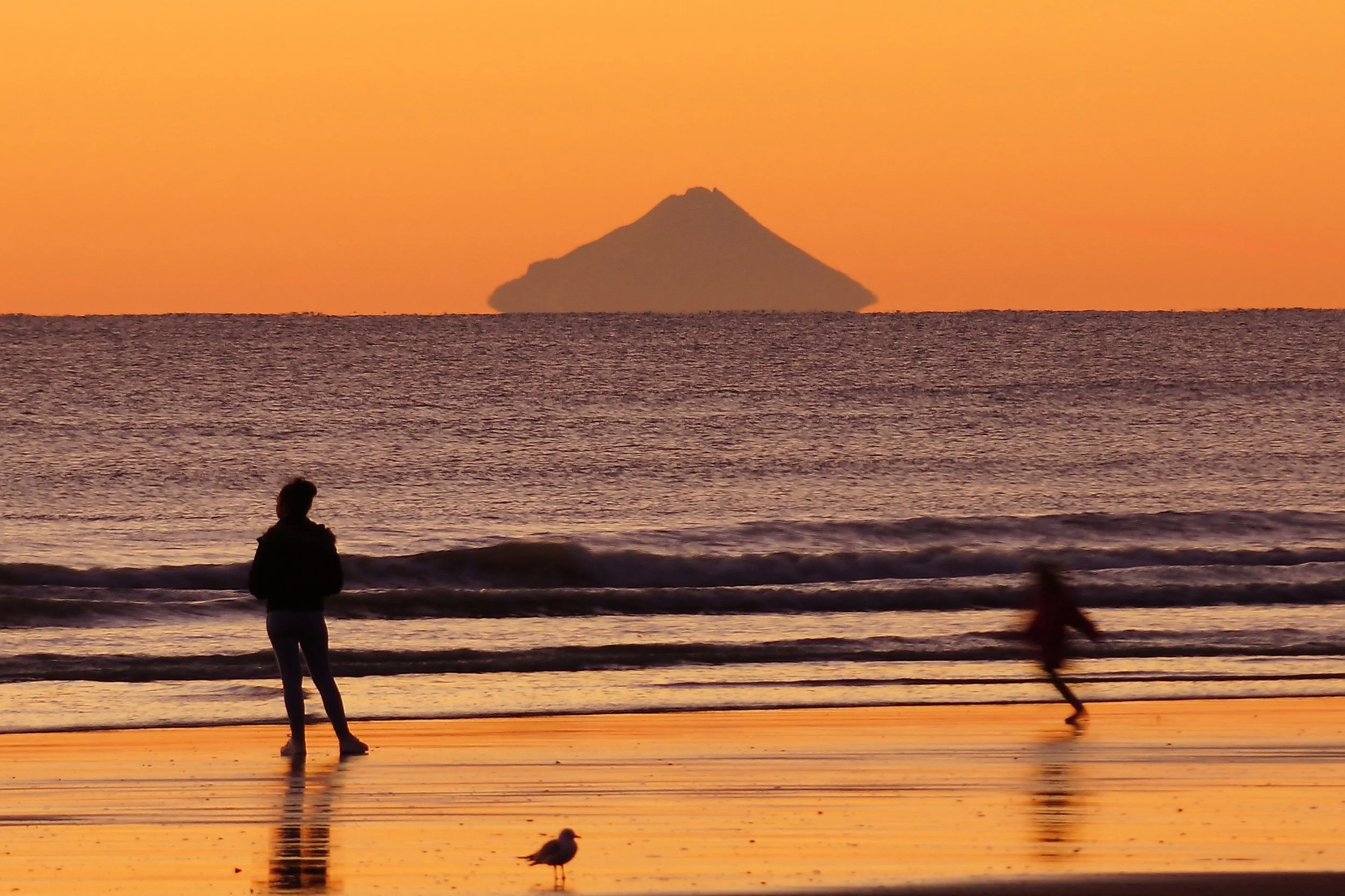 People walking and standing on a beach at sunset with the ocean in the background, and a mountain or volcano visible in the distance under an orange sky.