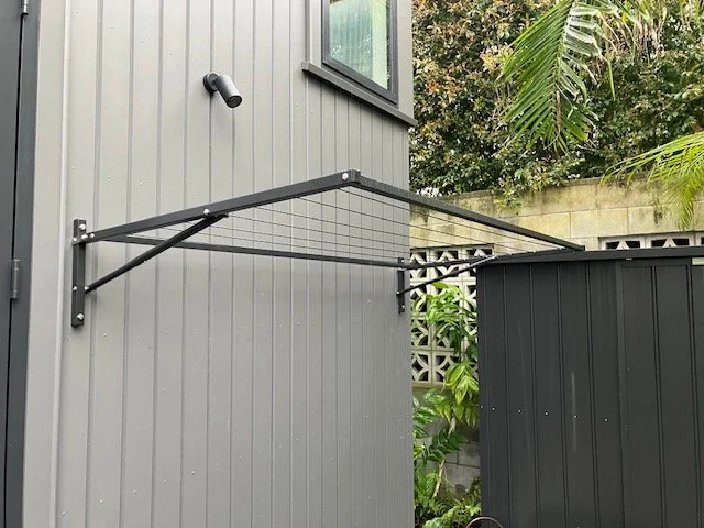 Black metal wall-mounted pull-up bar installed on the exterior gray siding of a building next to a black shed, with green foliage and trees in the background.