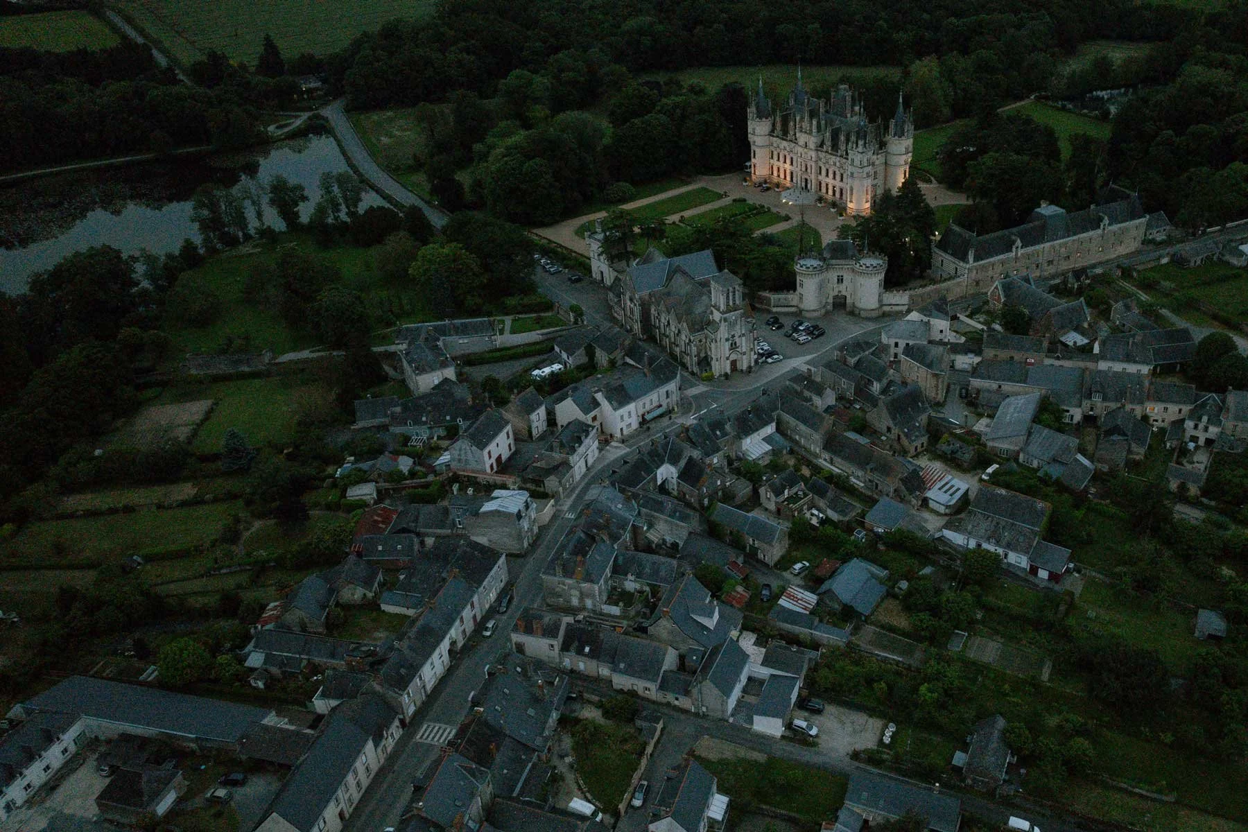 Aerial view of a South of France village with a large, illuminated castle surrounded by trees and quaint houses at dusk.