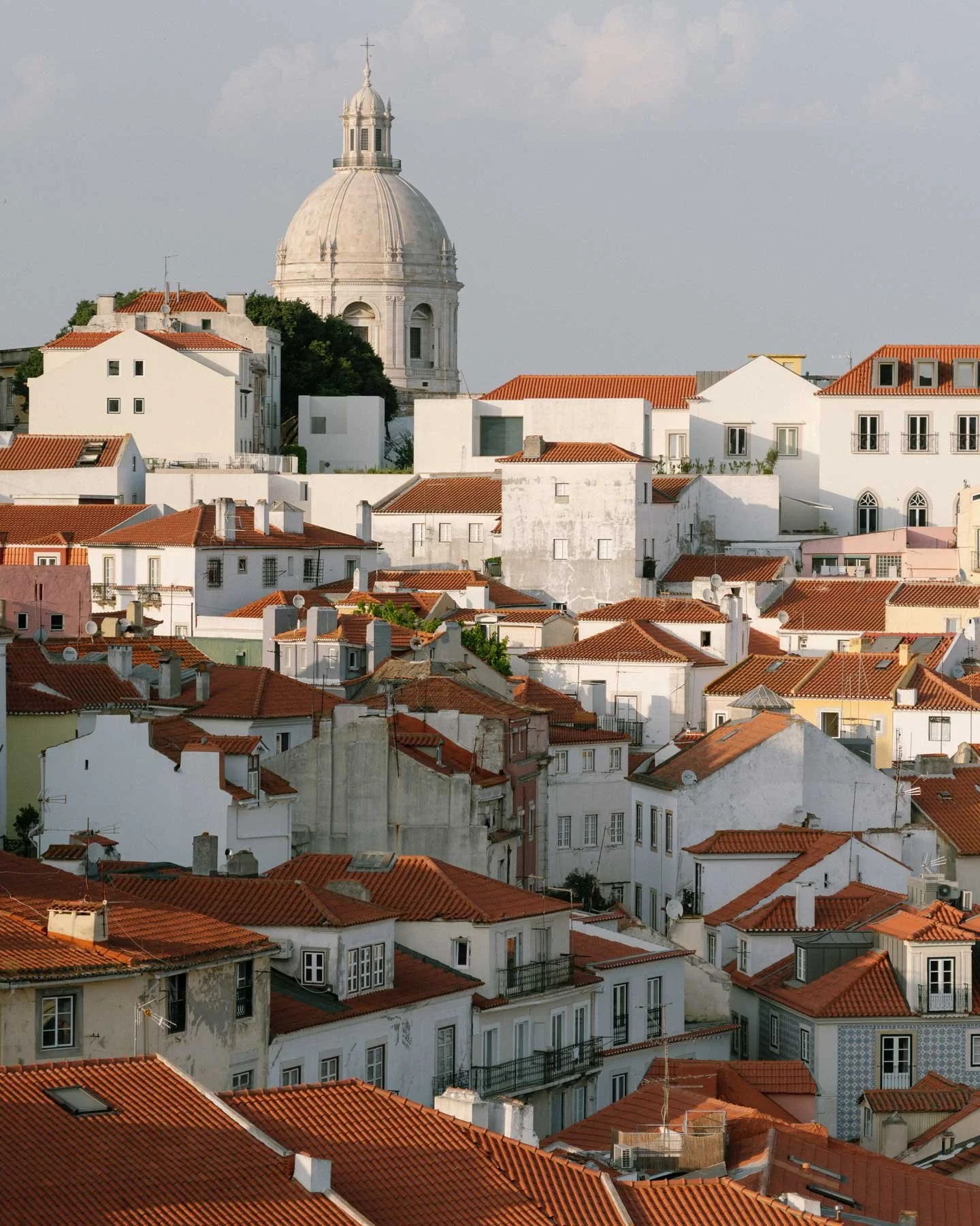 A cluster of white buildings with red-tiled roofs and a large domed church in the background under a cloudy sky.