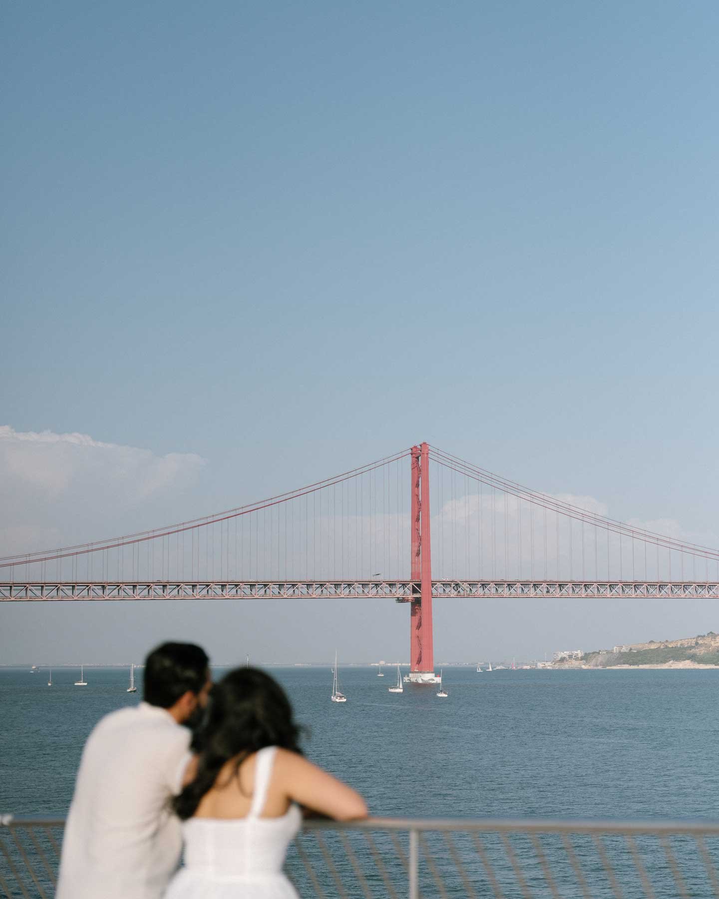 A couple leans on a railing, gazing at Portugal's iconic red suspension bridge over calm water—perfect for Indian weddings in Europe.