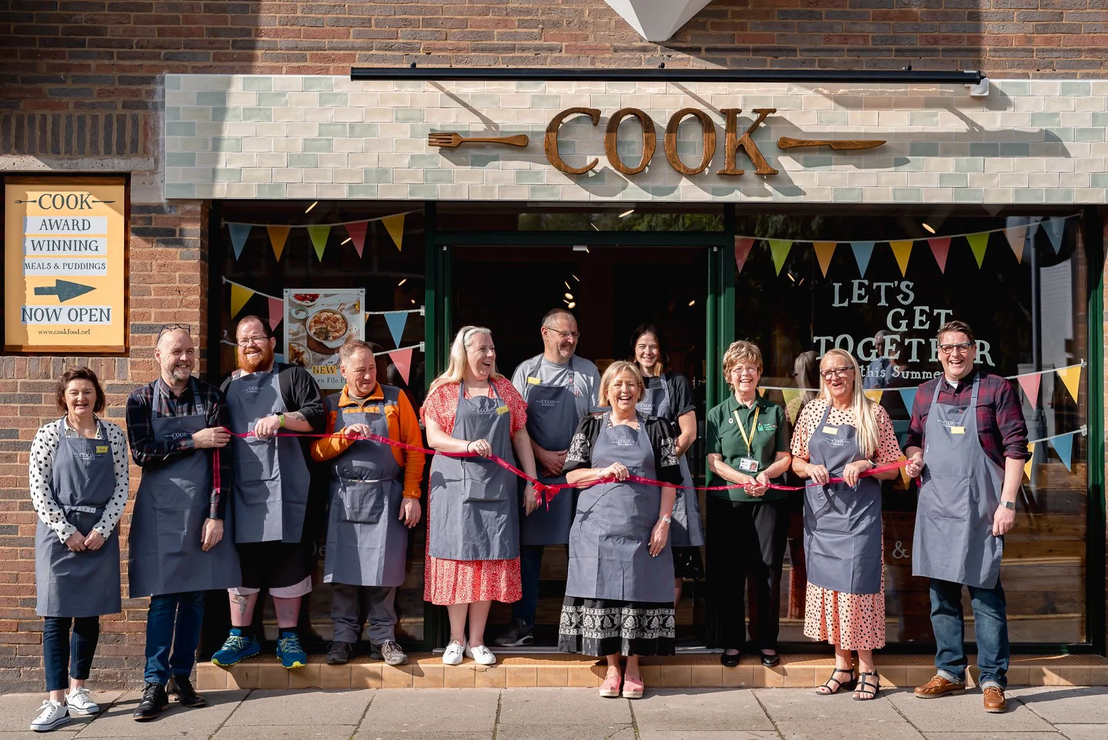 Group of people participating in a ribbon-cutting ceremony outside a restaurant named 'COOK' with a sign that reads 'Let's get together this summer' and colorful bunting decorations.