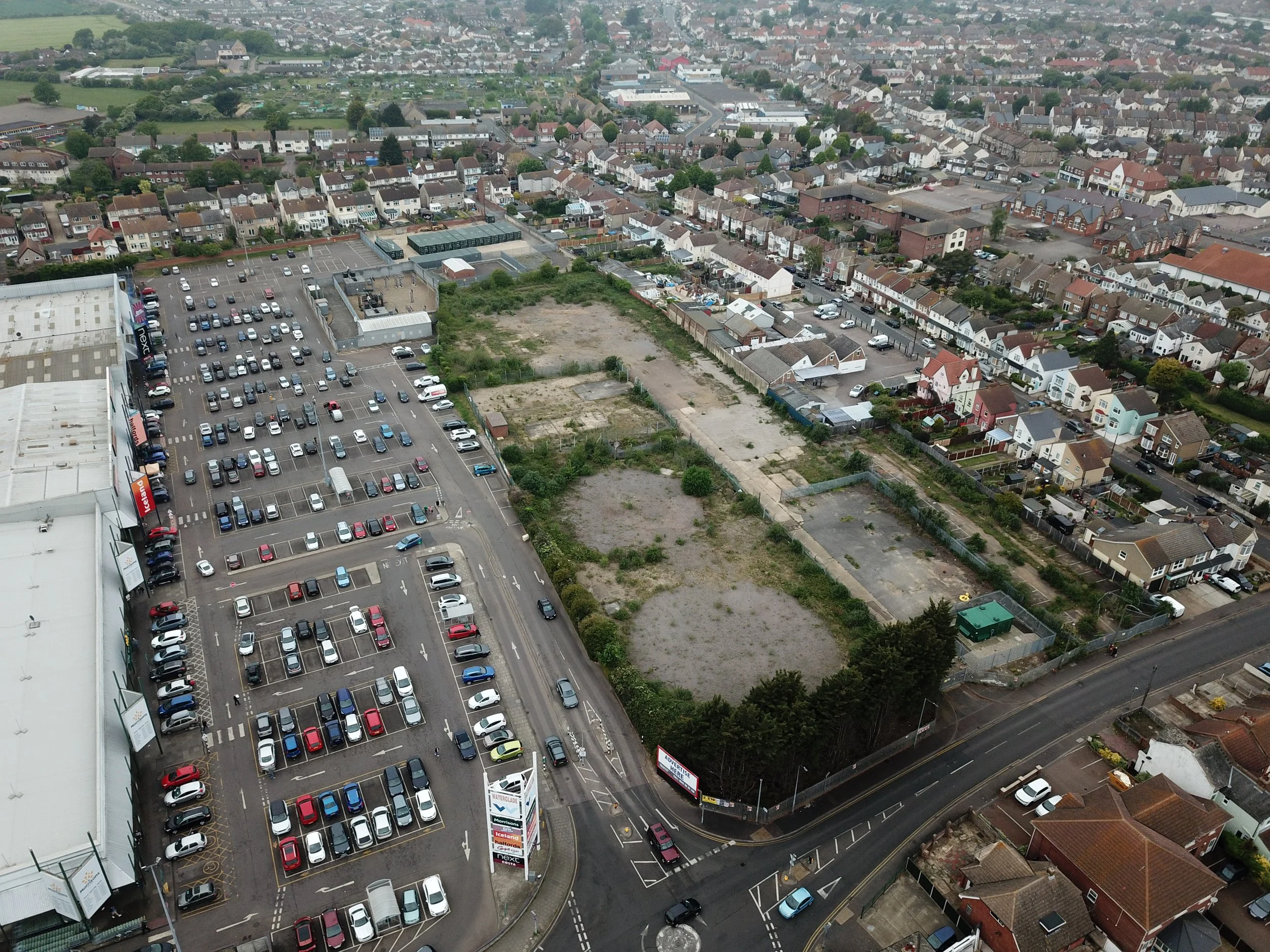 Aerial view of a large parking lot with many cars parked, adjacent to a shopping center or commercial building on the left side. Residential area with houses and small roads is visible on the right. There are some vacant lots with greenery in between