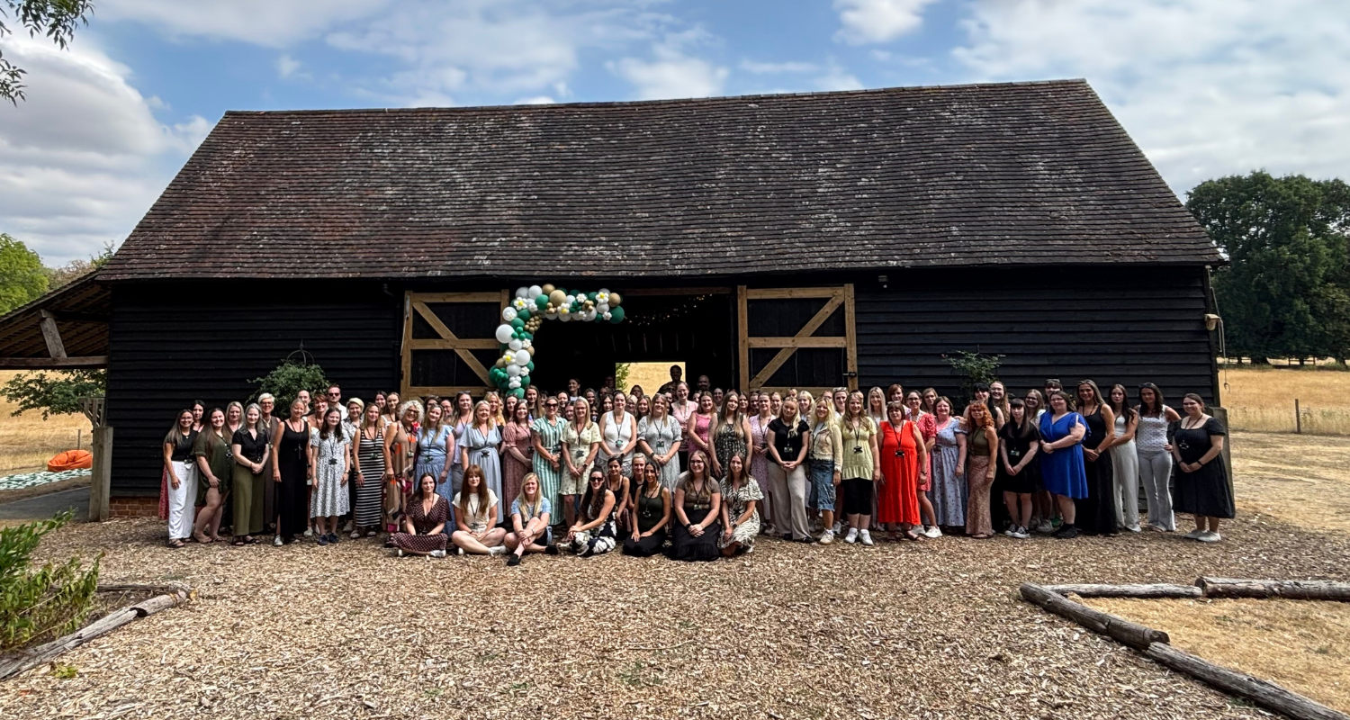 Large group of women gathered in front of a black barn decorated with green, white, and gold balloons for a celebration.