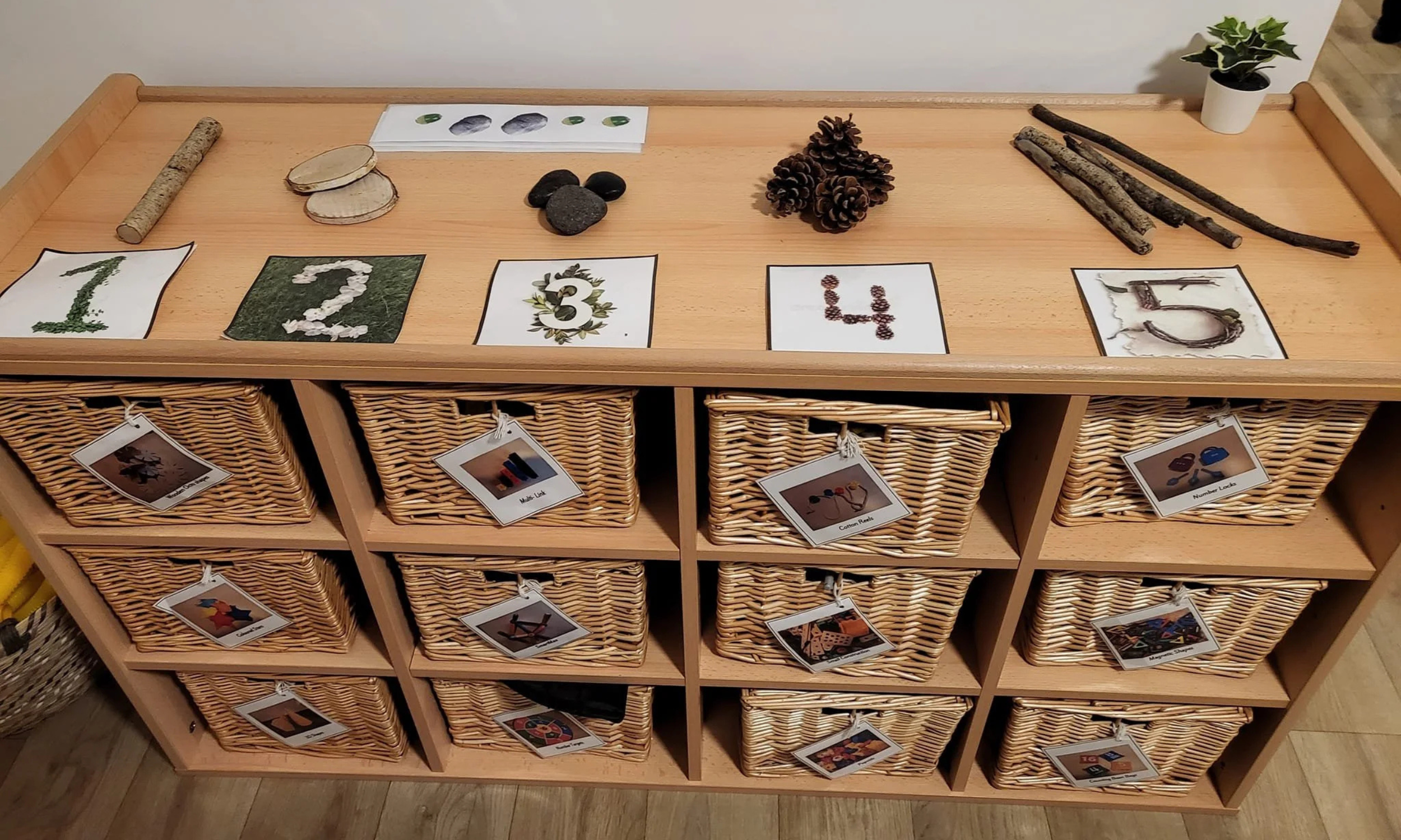 A wooden storage unit with wicker baskets, each with a label photo showing different items such as craft supplies and toys. The top surface displays natural objects like rocks, pinecones, sticks, and a potted plant.