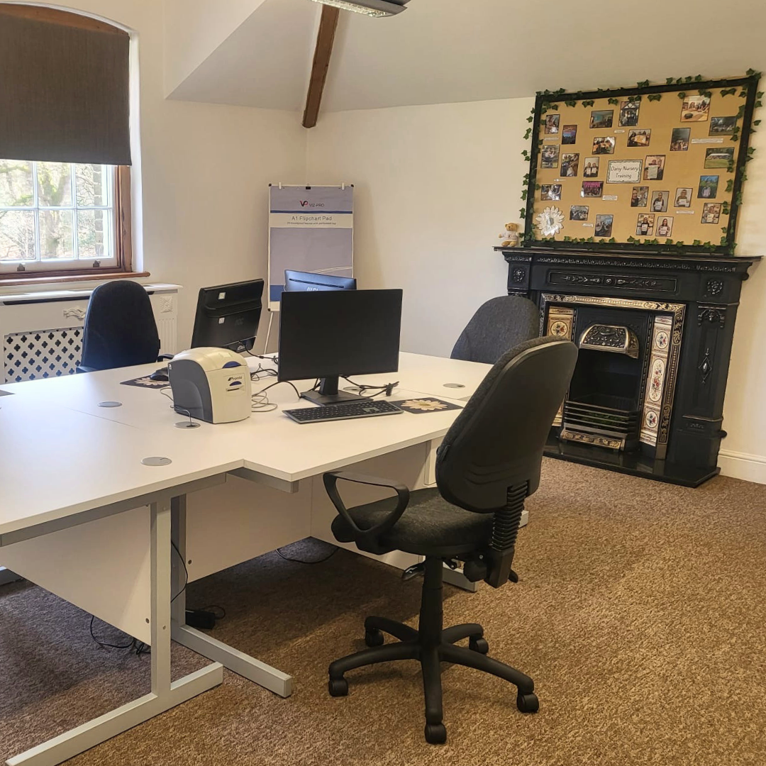 Office room with white desk, black office chair, computer monitors, a Victorian-style black fireplace, and a corkboard with photos.