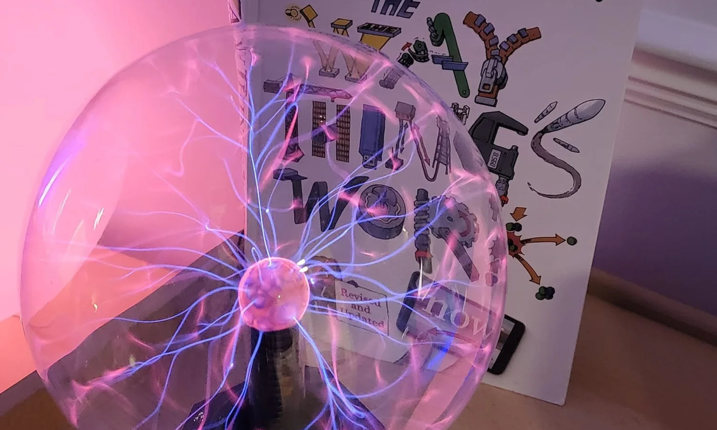 A plasma globe with electrical discharges inside, placed on a surface against a display showing educational science illustrations.