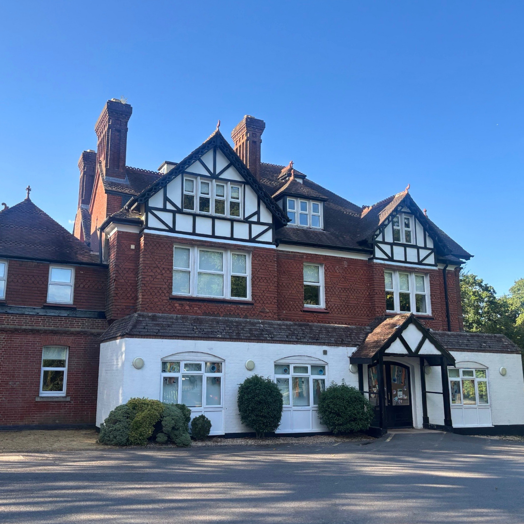 A large multi-story house with Tudor-style architecture, featuring red brick, white stucco with black timber framing, multiple windows, and a prominent front entrance with a black canopy, surrounded by shrubbery, under a clear blue sky.