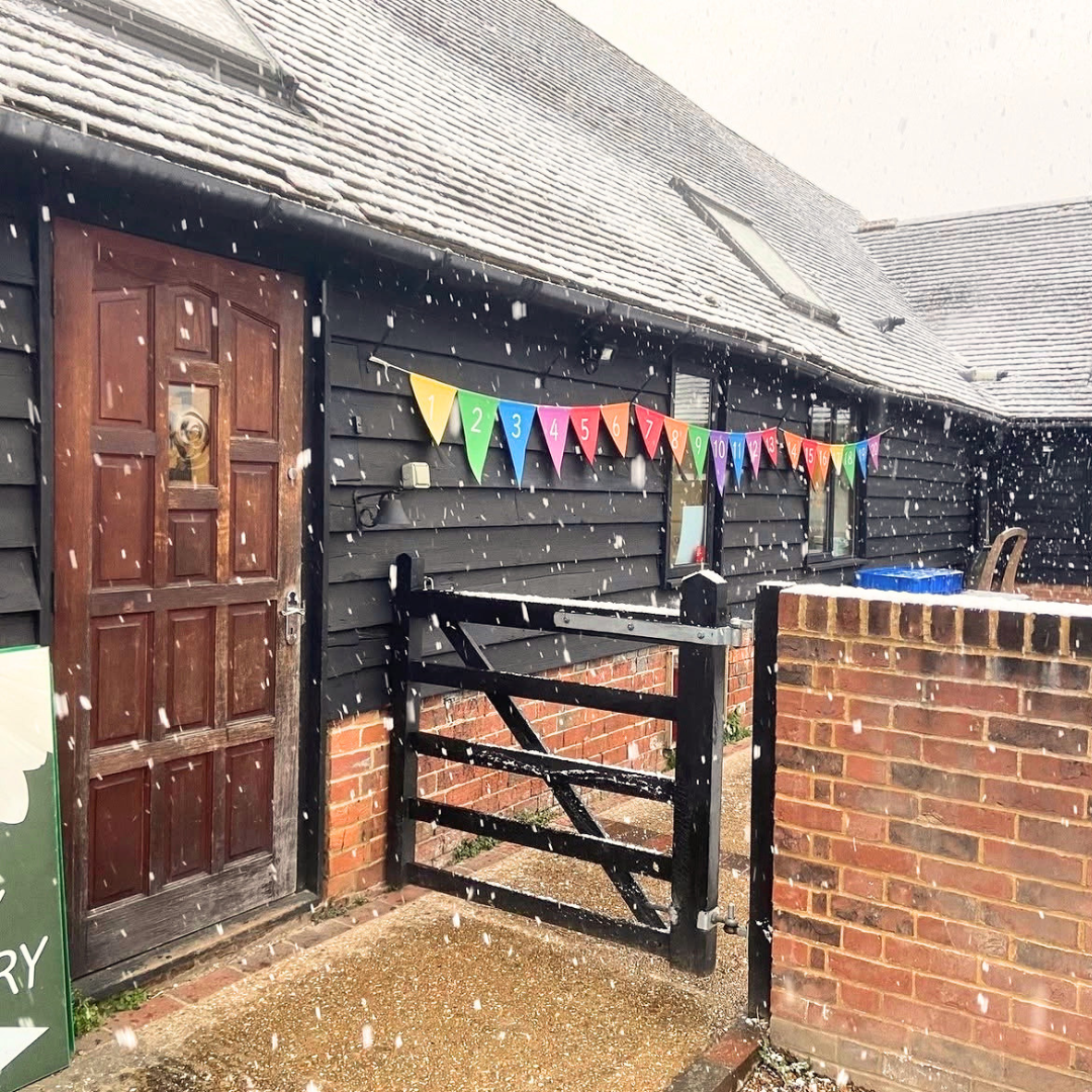 Snow falling outside a black wooden house with a brown door and a rainbow-colored number banner, with a brick wall and gate in front.