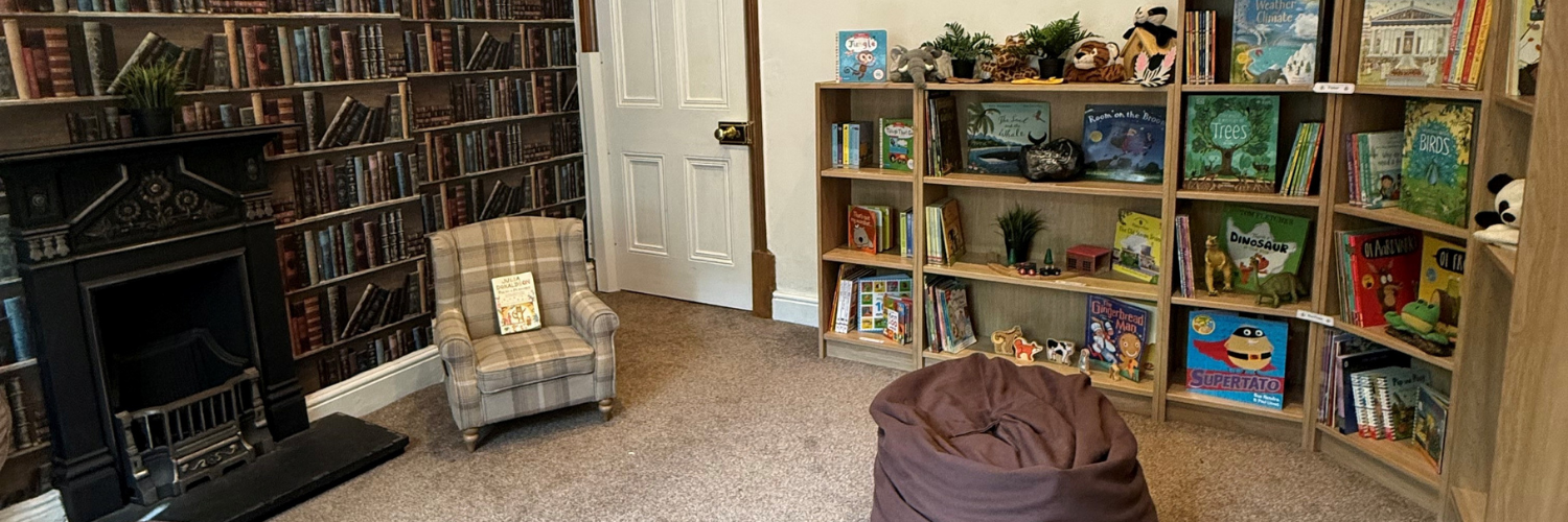 A cozy children's reading room featuring a checked armchair with a children's book on top, a large brown bean bag, and bookshelves filled with children's books, toys, and plush animals. There is a fireplace with a decorative wood mantel and a bookshelf with a wallpaper-like pattern resembling a library wall. The room has a carpeted floor and a white door.
