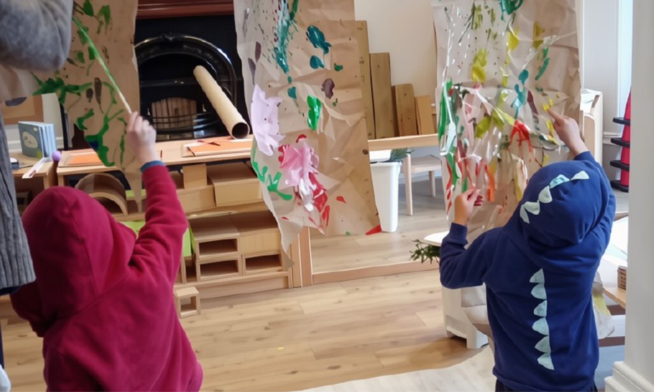 Children wearing hooded jackets, one in pink and one in blue, painting on large sheets of paper attached to a wooden frame in a classroom setting.