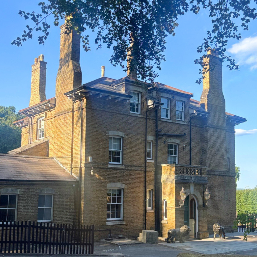 A historic brick building with multiple chimneys, arched windows, and decorative stonework, featuring two lion statues at the entrance and a black metal fence in front.