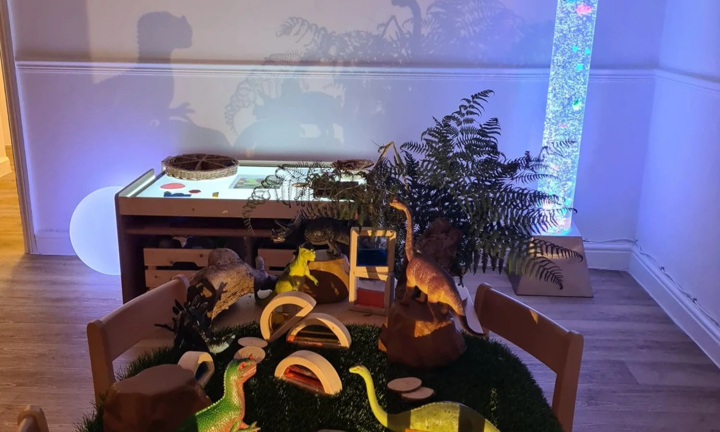 Indoor scene with children’s toys and dinosaur figurines on a carpeted floor, a table with a display and plants, illuminated by colorful blue lighting.