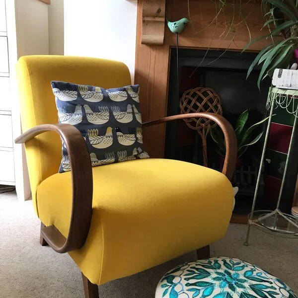 Yellow upholstered armchair with wooden armrests and a decorative pillow with a leaf pattern, in a cozy living room setting with a fireplace, plants, and a patterned footstool.