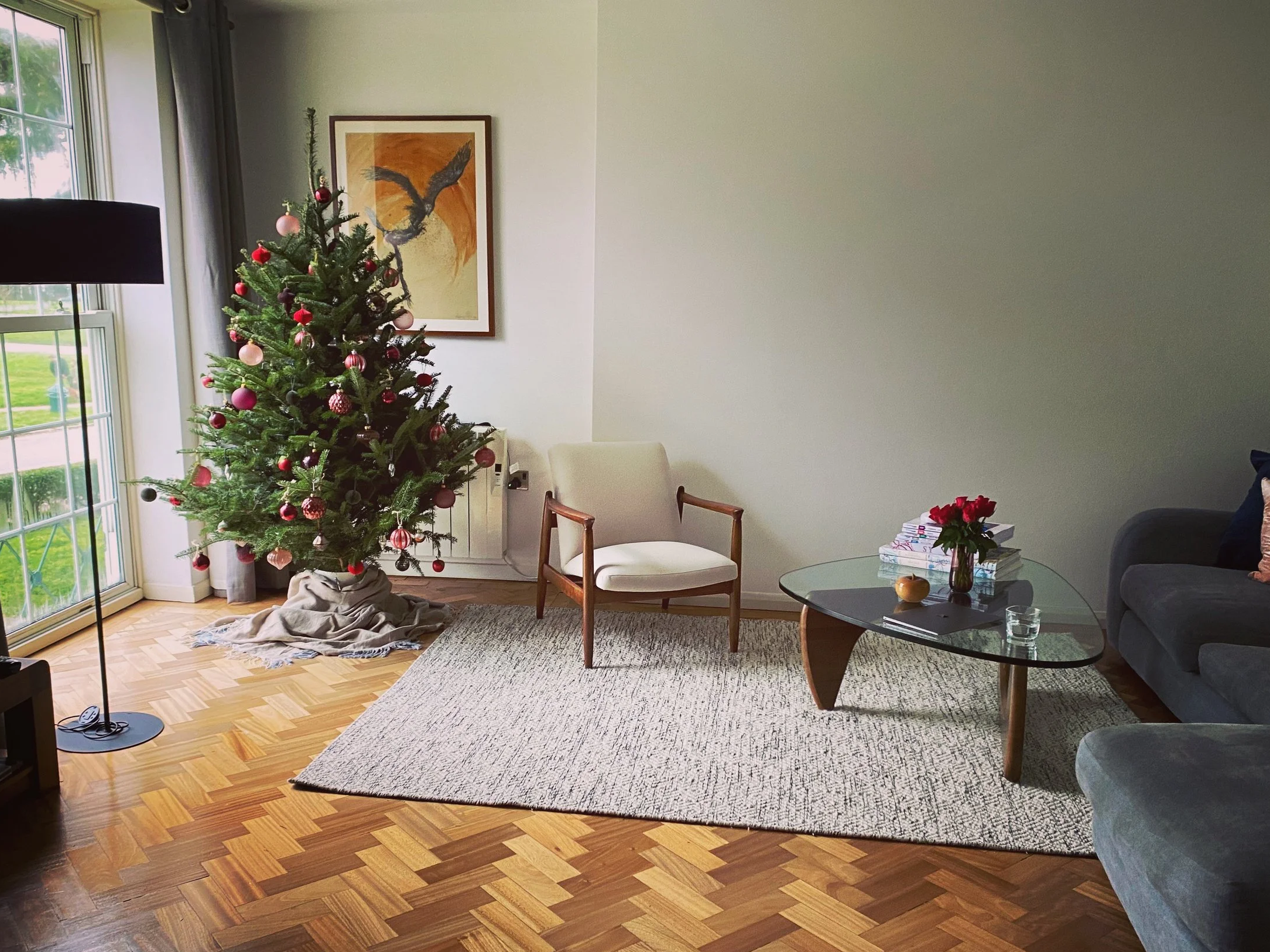Living room decorated for Christmas with a Christmas tree, positioned near a large window. There are mid-century chair, a gray sofa, a glass coffee table with a stack of books, red flowers, and a painting of a bird on the wall.