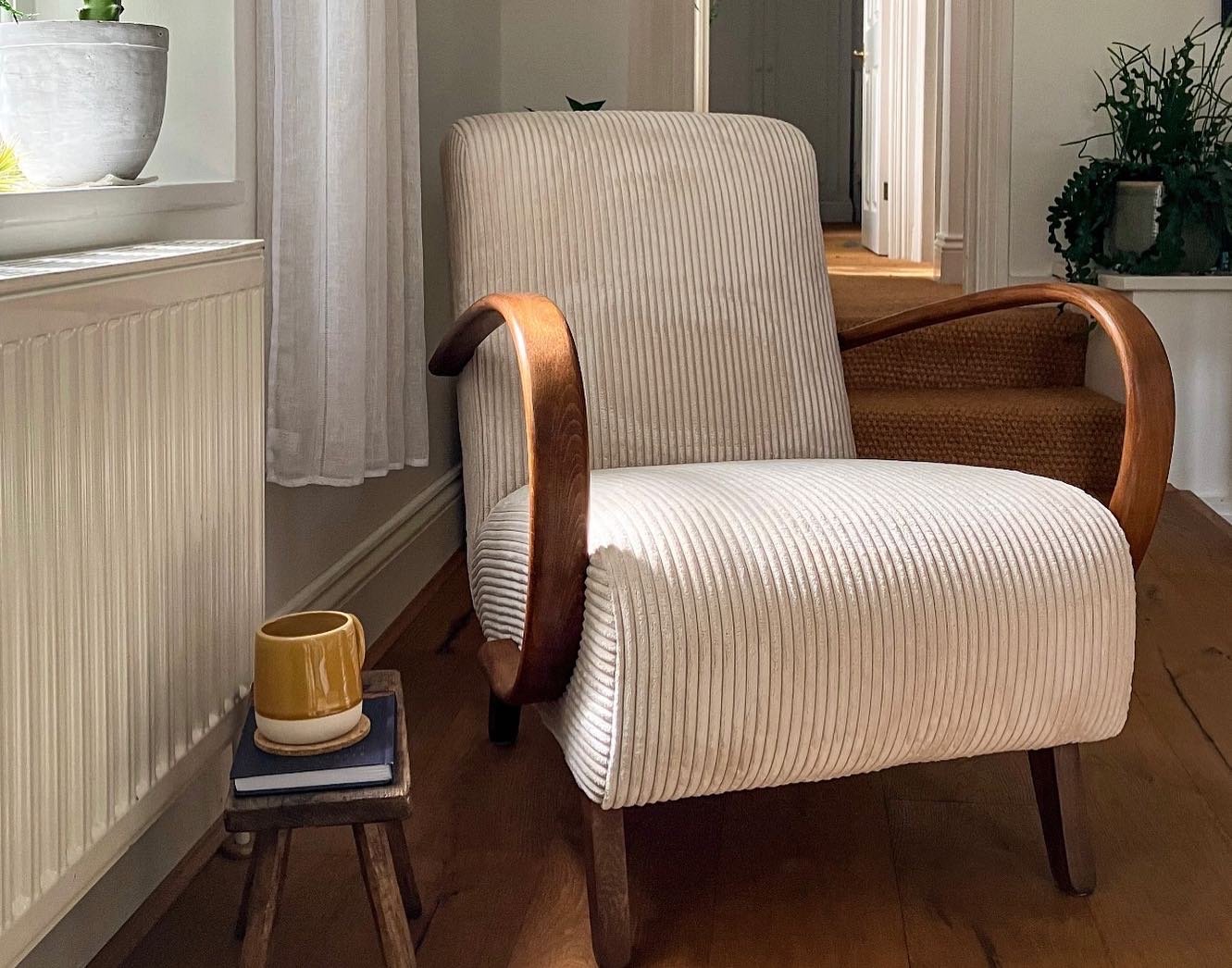 A white cream art deco armchair with wooden armrests and legs, positioned beside a small wooden side table with a ceramic mug and a closed book on it, in a cozy scandinavian style or japandi room with hardwood floors and neutral-coloured walls.