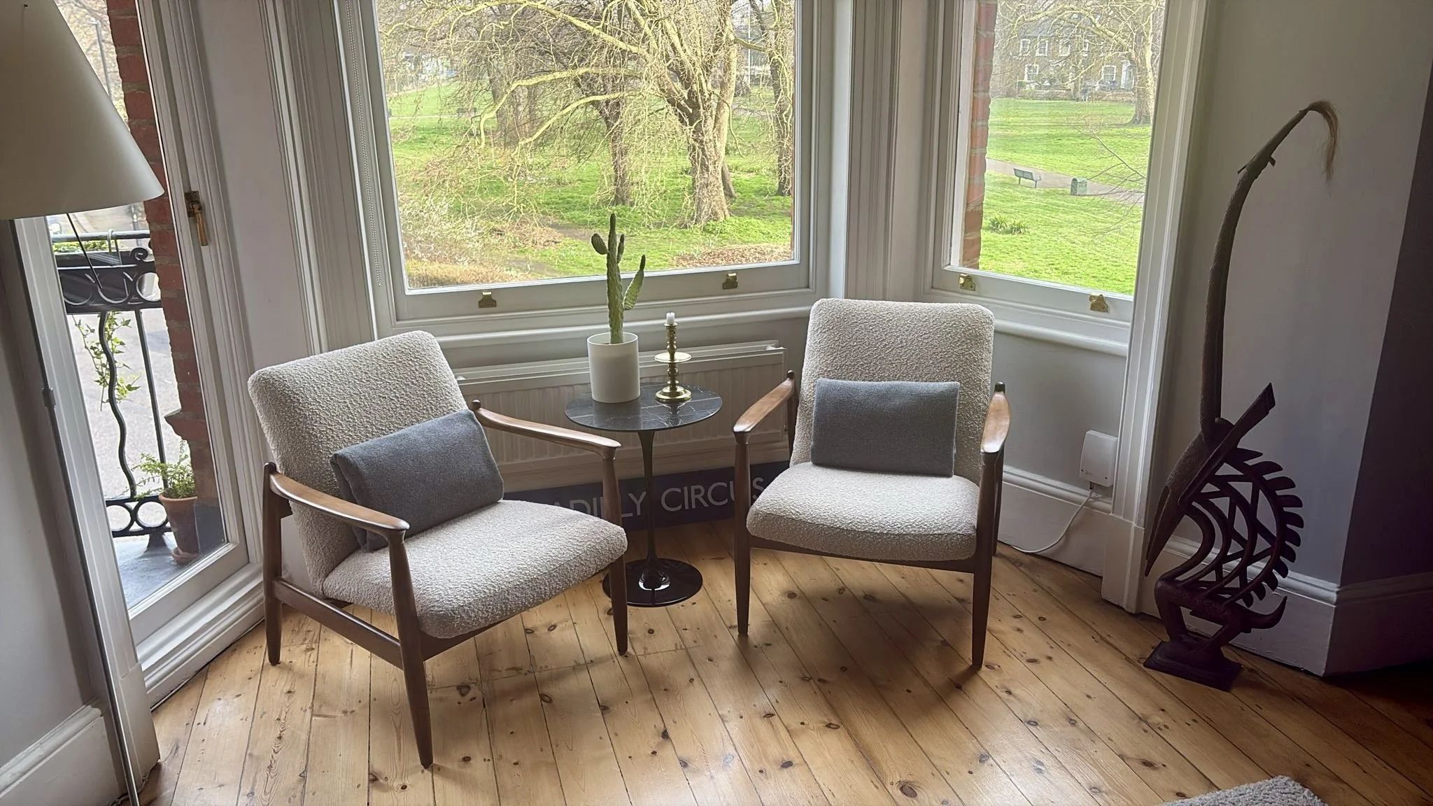 A cozy corner inside a house with two mid-century vintage armchairs upholstered in white boucle, at a small round black table, which has a potted cactus and a candle holder. Large windows overlook a green outdoor area with trees and walking paths.