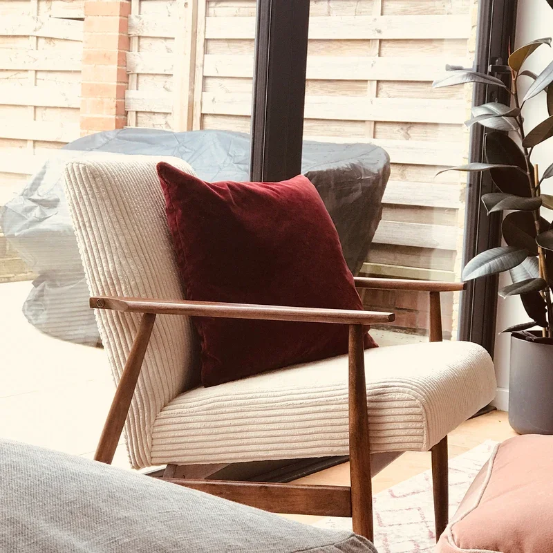 A cozy white mid-century armchair with wooden armrests and a cream corduroy fabric, with a dark red velvet pillow, near large glass doors leading to a backyard with a wooden fence and outdoor furniture.