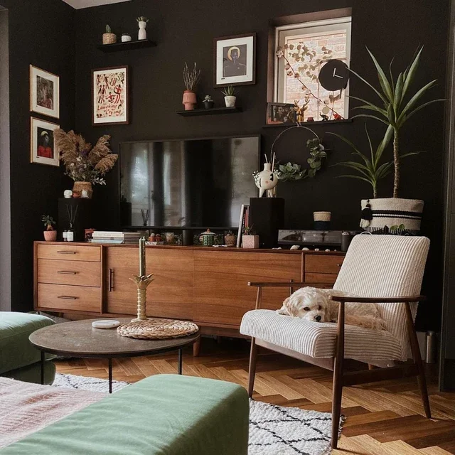 Cozy living room with black accent wall, wooden TV console, framed art, plants, and a white vintage mid-century lounge chair where a dog is resting, adjacent to a green sofa and a round coffee table.