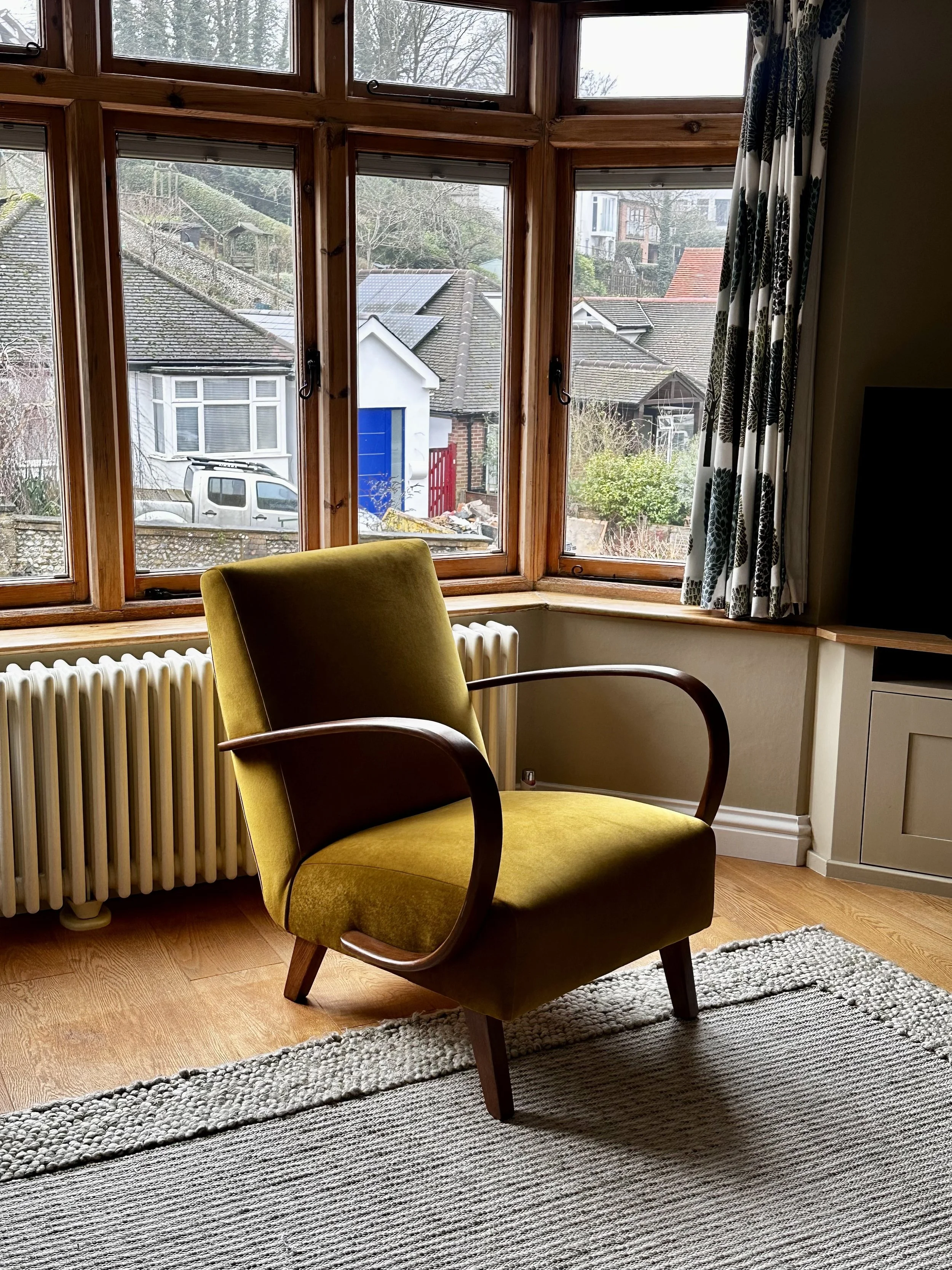 A vintage mustard yellow armchair with dark wooden armrests and legs, positioned in front of a large bay window with wooden frames, overlooking a residential neighborhood. There is a radiator below the window, a patterned curtain on the right, and a textured gray rug on the wooden floor.
