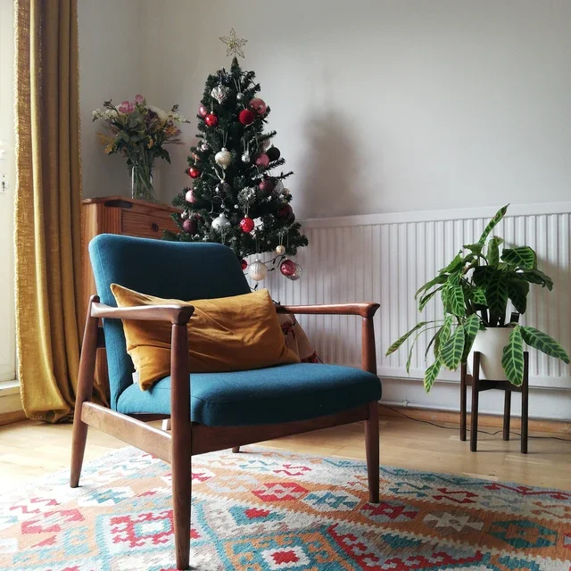A decorated Christmas tree with ornaments and a star topper, a wooden side table with a vase of flowers, a mid-century modern blue armchair with a yellow pillow, a potted plant, a colorful area rug, and a window with yellow curtains in a cozy living room.