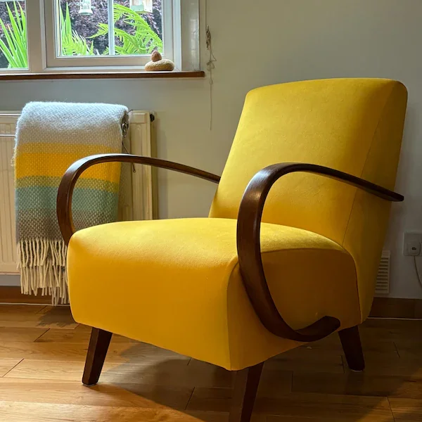 Art deco yellow velvet armchair with curved wooden armrests in a cozy indoor setting, with a window showing greenery outside, a colourful blanket draped over a radiator, and a small decoration on the windowsill.