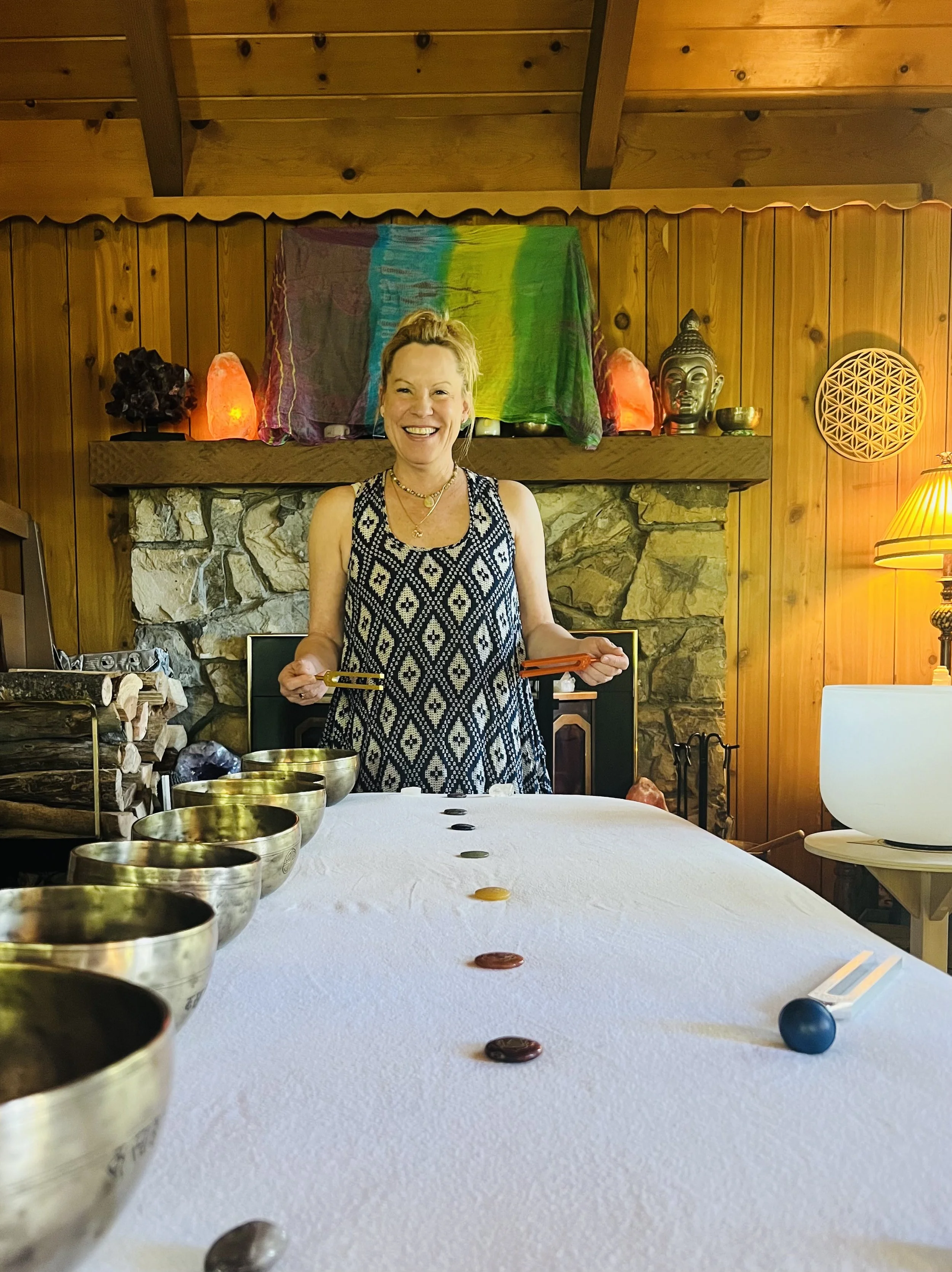 Rachelle Bellici smiling and standing behind a massage table with singing bowls, holding tuning forks, in a cozy wooden room with spiritual decor.