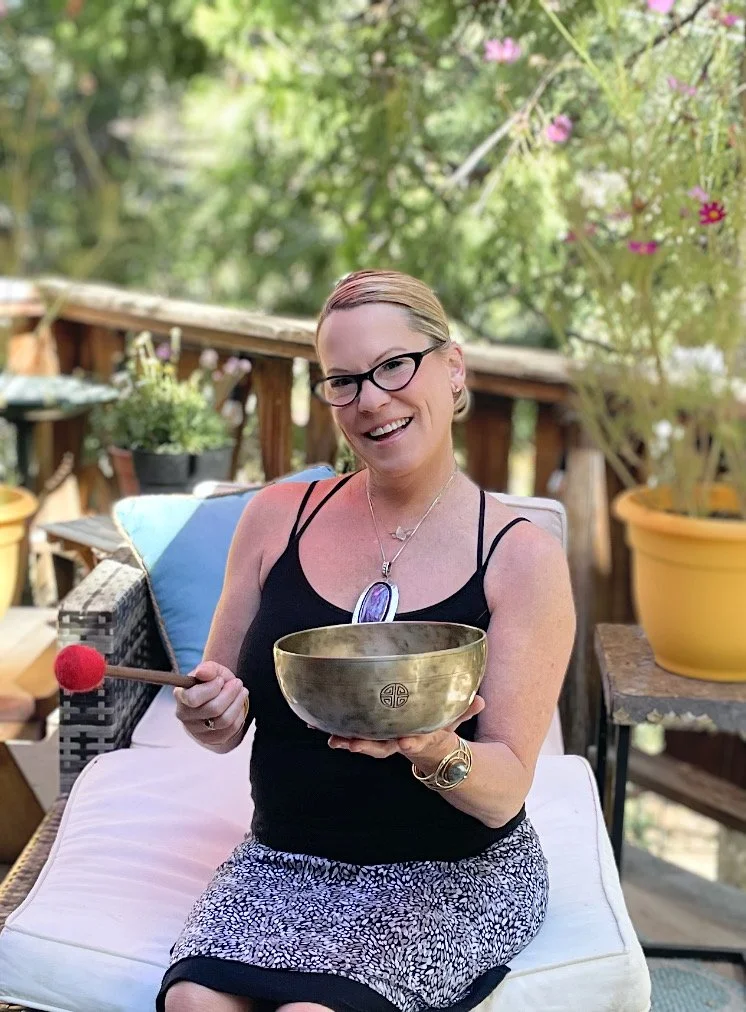 Rachelle Bellici, sound frequency healer, sitting outside, smiling, striking a metal singing bowl