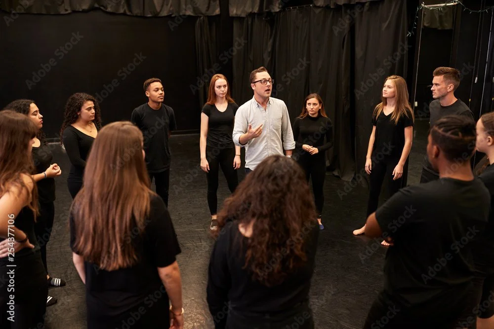 A man leading a rehearsal or acting class with a diverse group of students standing in a circle in a theatre studio.