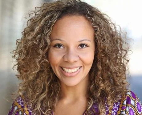 Close-up of a smiling woman with curly hair, wearing a colorful patterned top.