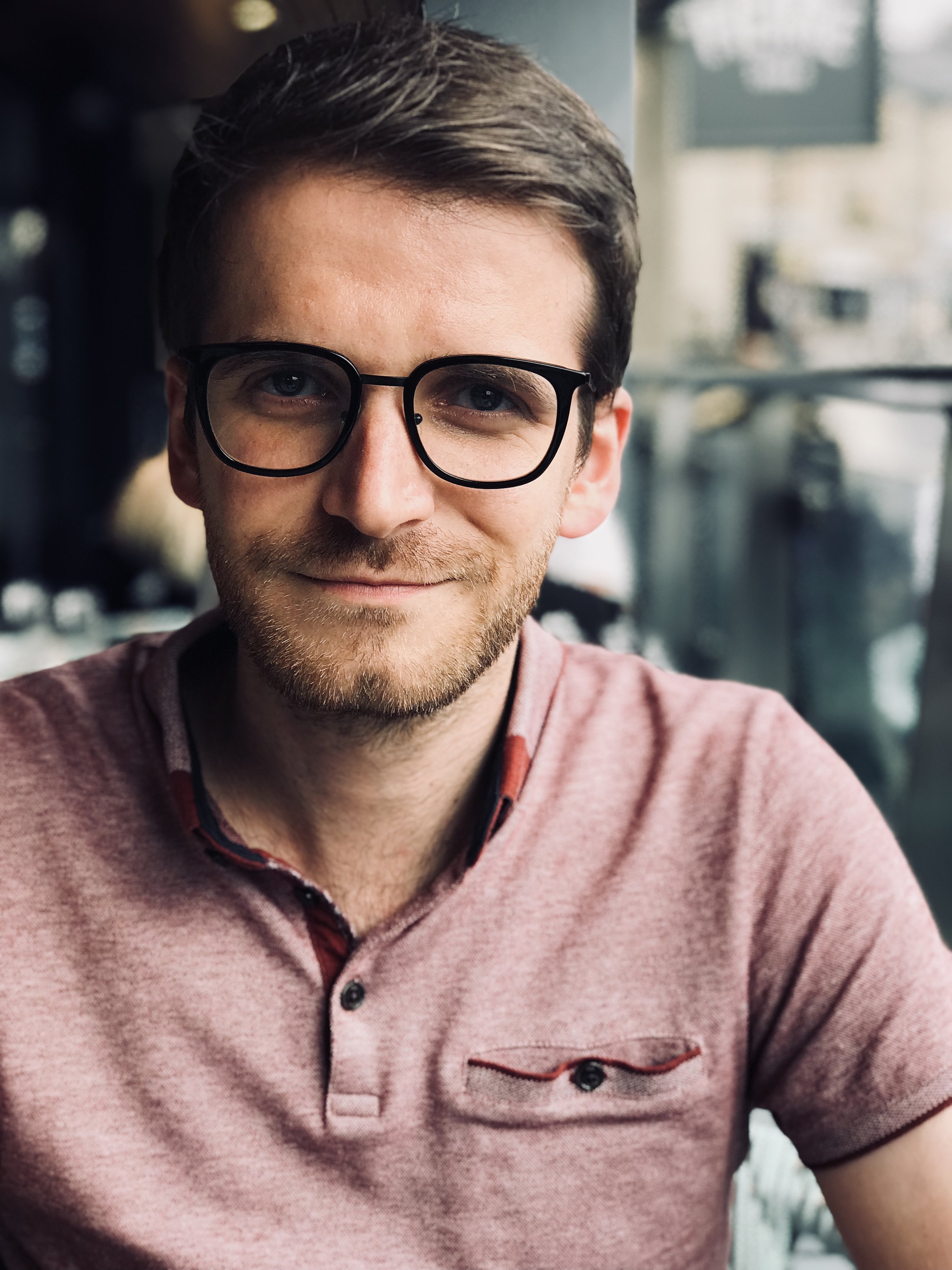 A young man with glasses and a light beard smiling in a cafe.