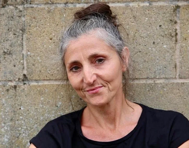 A middle-aged woman with gray hair styled in a bun, wearing a black top, smiling softly, standing against a brick wall.