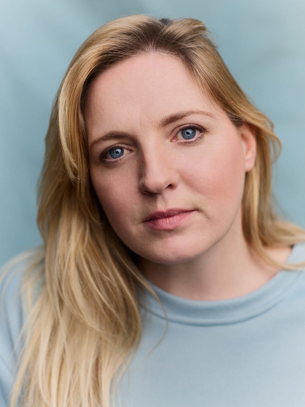 Close-up portrait of a young woman with blonde hair and blue eyes against a light blue background.