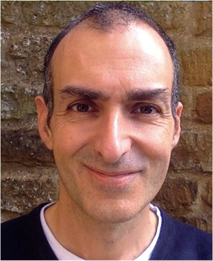 Close-up of a man with short dark hair, smiling, standing in front of a brick wall.