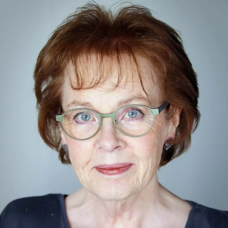 Close-up portrait of an elderly woman with short, reddish-brown hair, wearing round glasses with light green frames, and a dark top, against a neutral background.