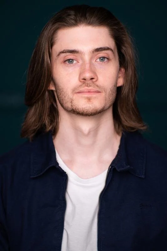 Portrait of a young man with long brown hair, light skin, and a slight beard, wearing a navy blue jacket over a white t-shirt, against a dark background.