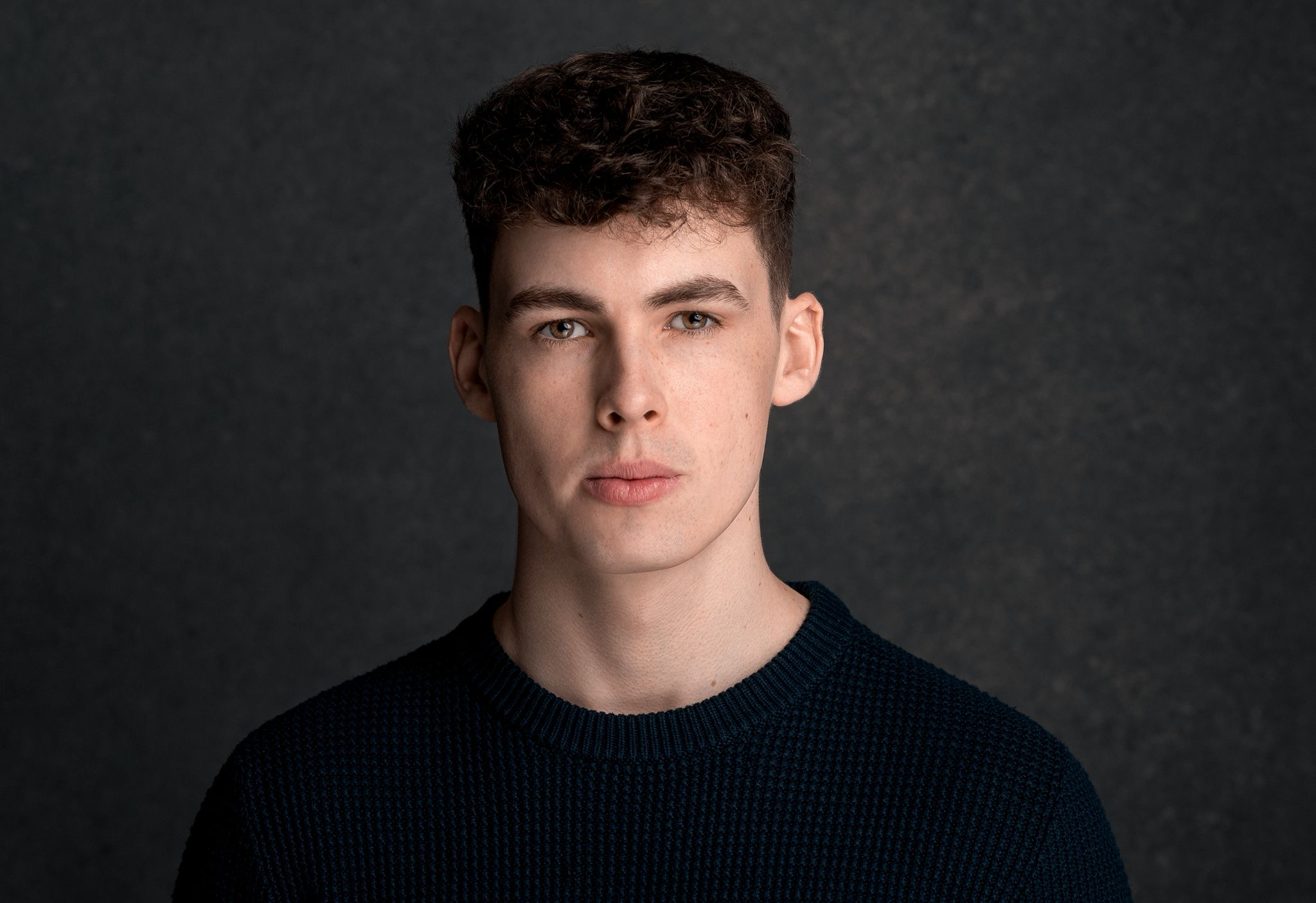 Portrait of a young man with short, curly dark hair and fair skin, wearing a dark sweater, against a dark background.