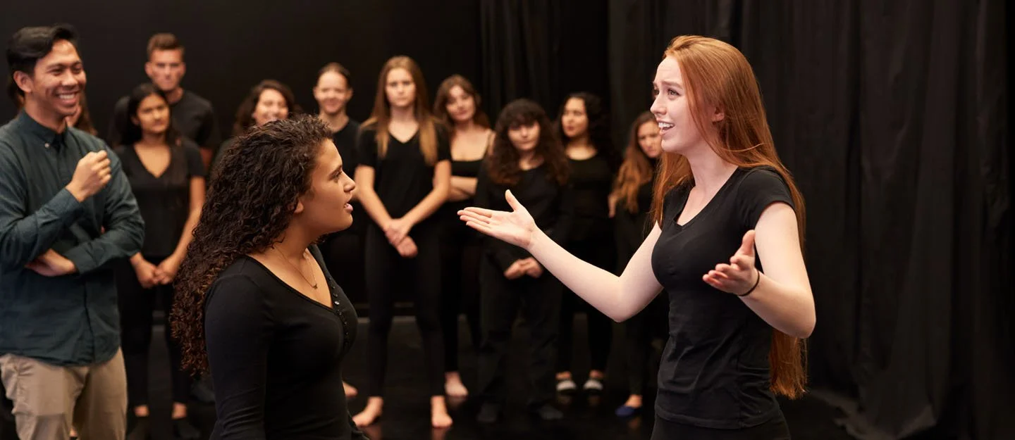 Two women with dark clothing engaged in a conversation or practice, surrounded by a group of women and men watching in a black room or stage.