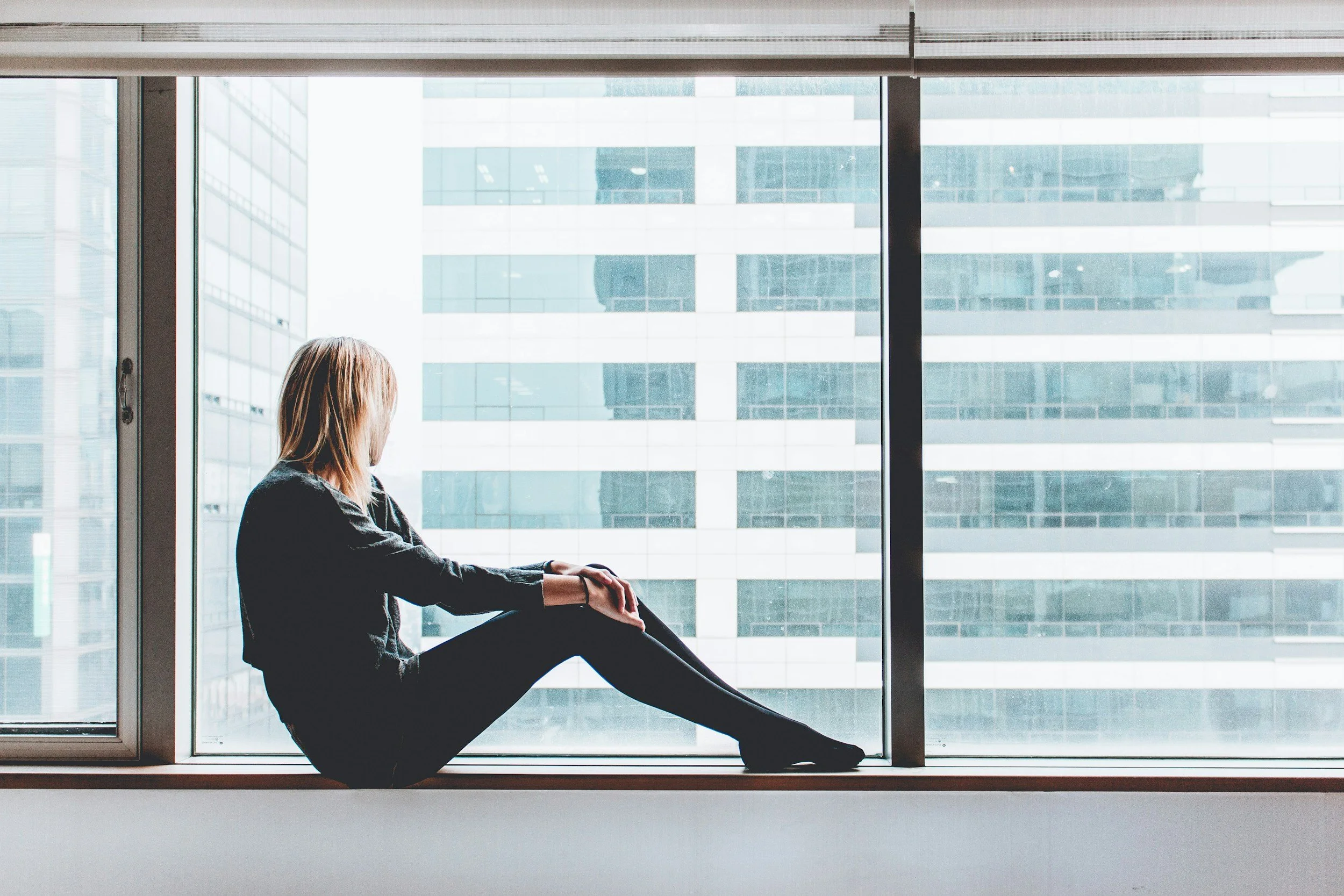 Eine Frau sitzt nachdenklich am Fenster in einem modernen Büro mit Blick auf eine Gebäudenfassade aus Glas.