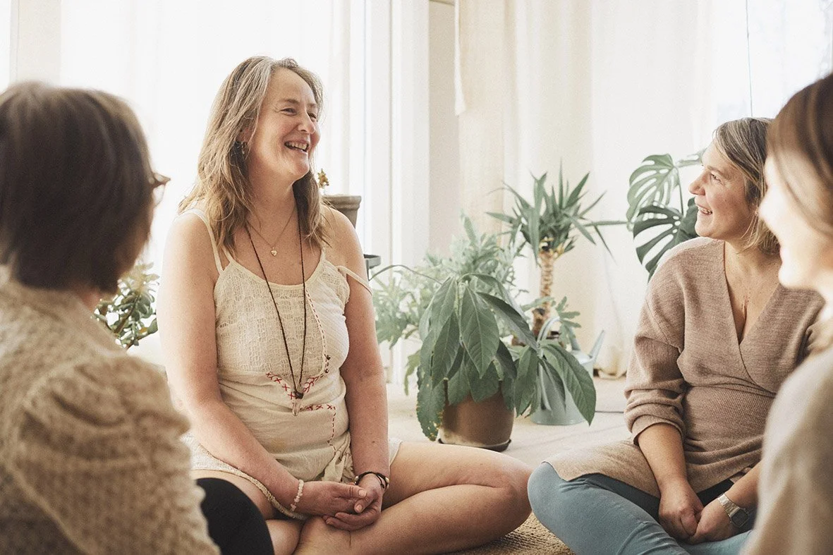 Four women sitting on the floor in a circle, smiling and laughing, with plants in the background.
