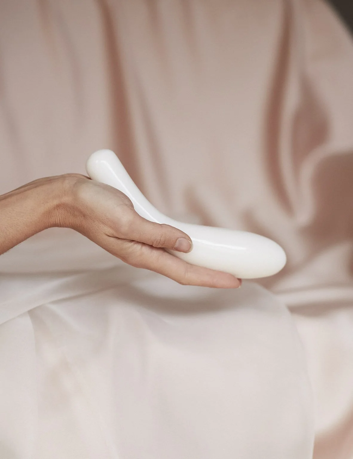 Close-up of a person's hand holding a smooth, white, curved object, with a soft pink fabric background.