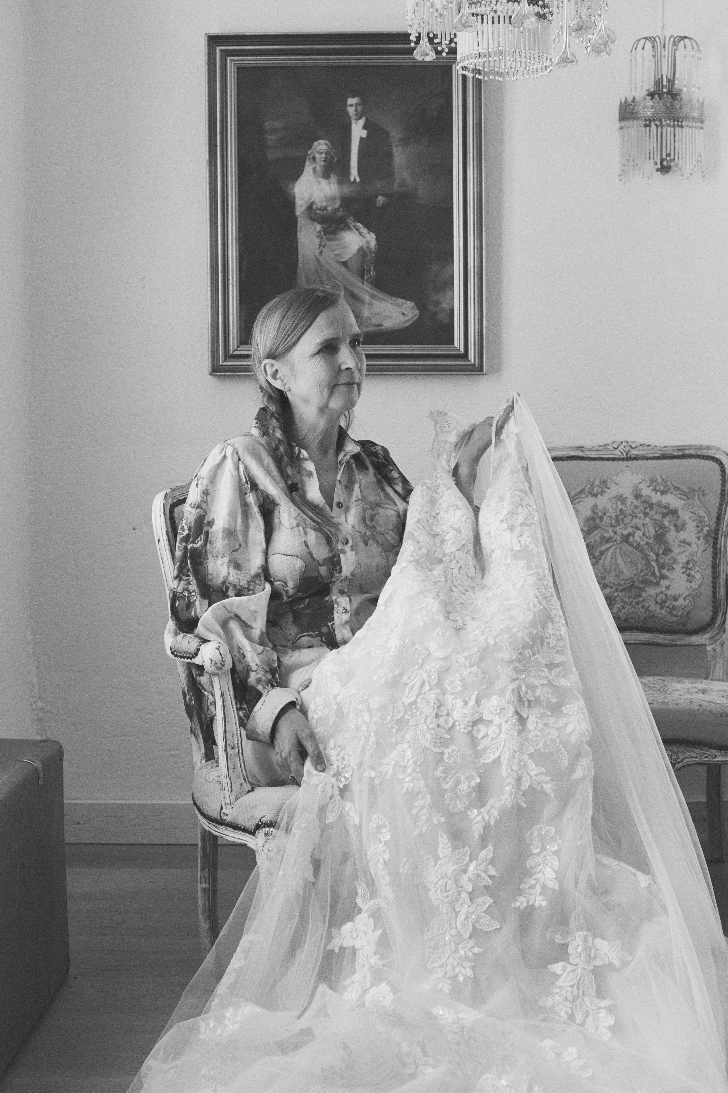 An elderly woman sitting on an ornate chair, holding a wedding dress, in a room with a large portrait of a young couple on the wall and chandeliers hanging from the ceiling.