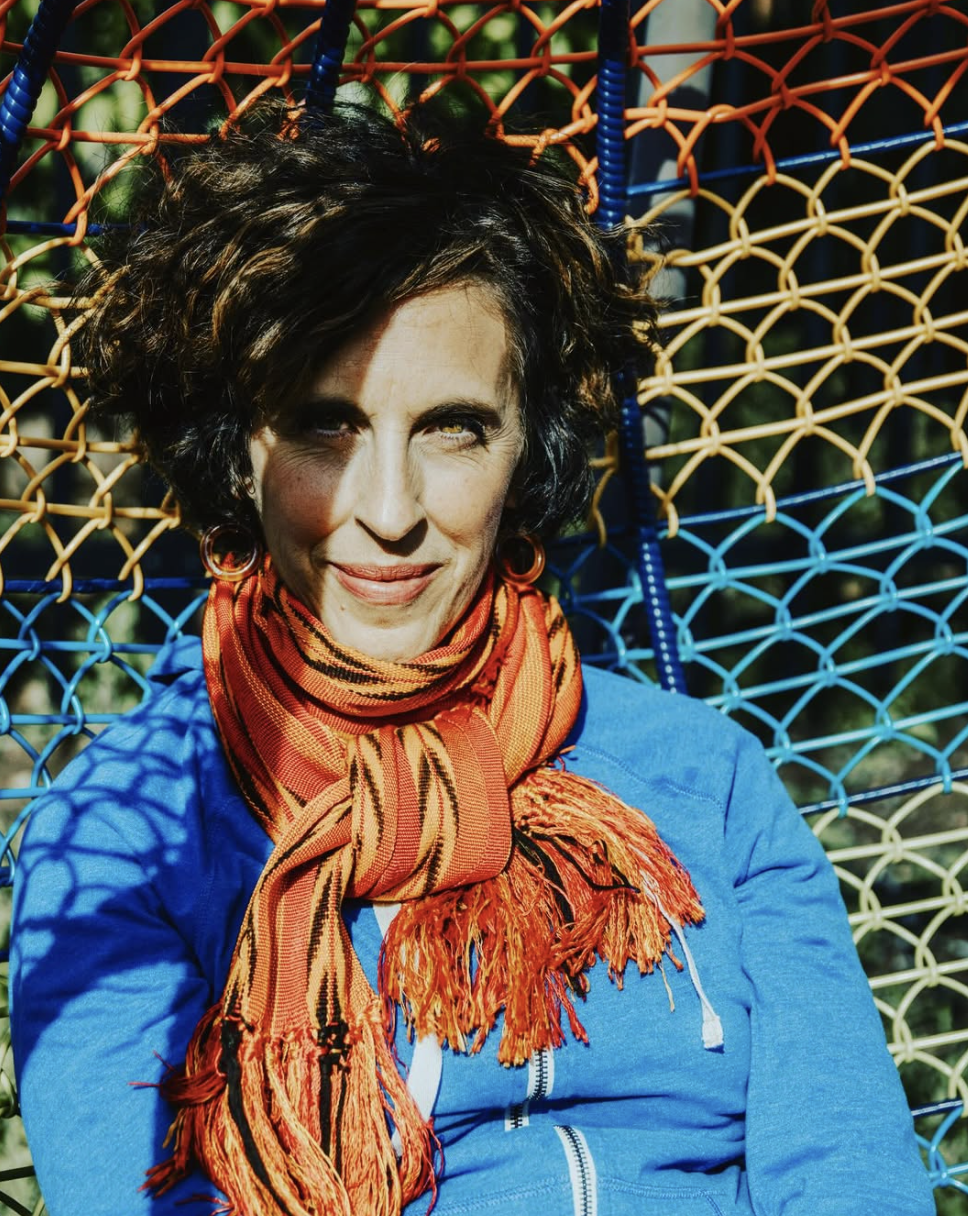 A woman with short, curly dark hair and light skin, wearing a bright blue jacket and an orange striped scarf, sitting against a colorful woven chair outdoors.