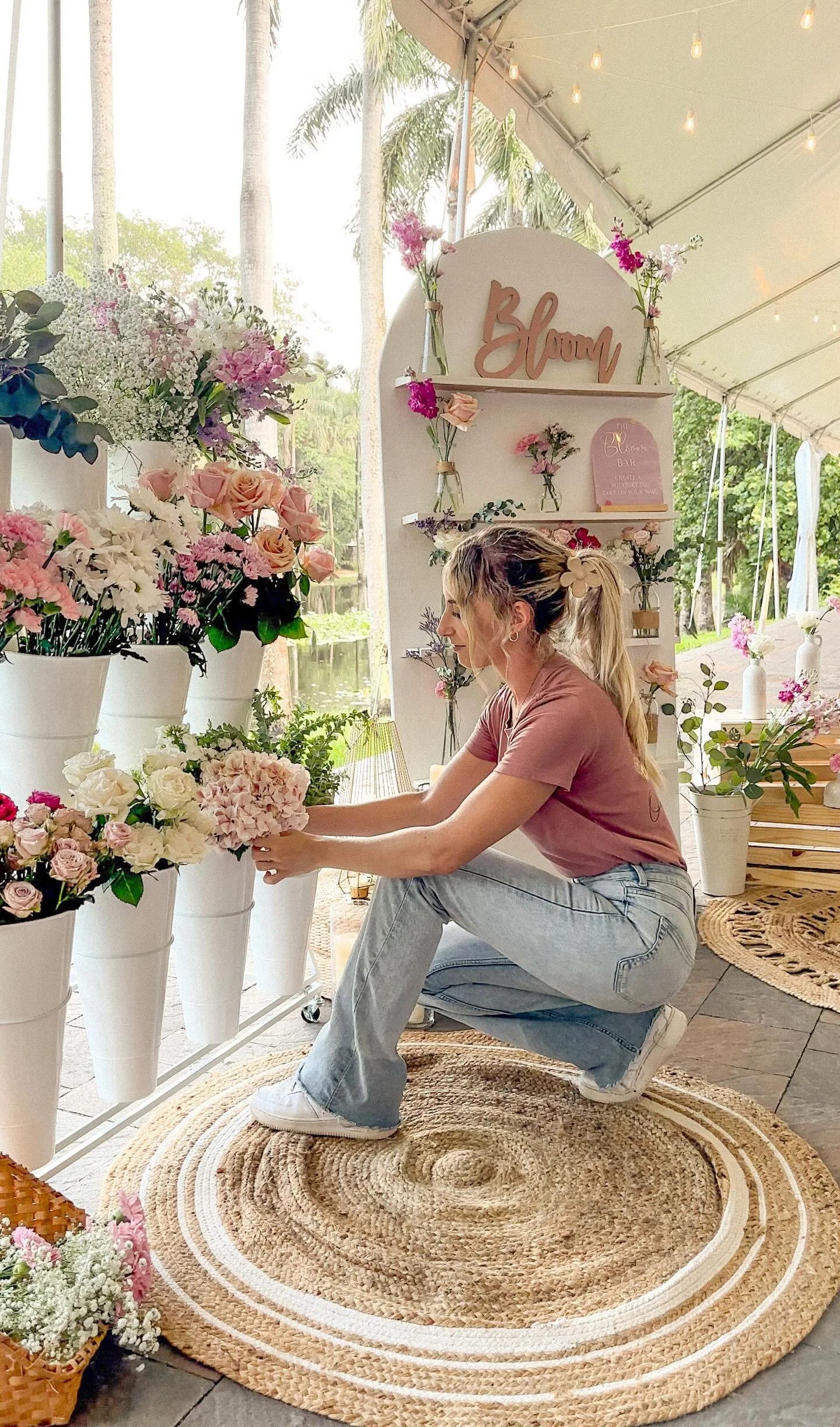 Woman arranging flowers in a tent with floral decorations, vases, and a "Bloom" sign.