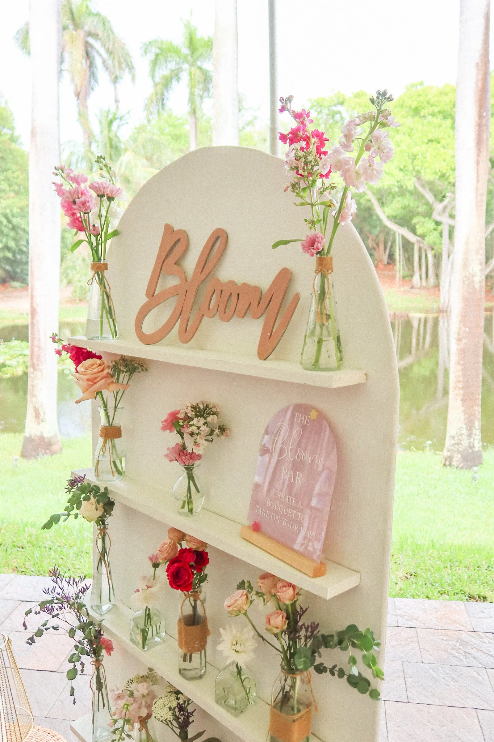 Flower bar with "Bloom" sign, featuring various flowers in vases on shelves, set in a garden setting.