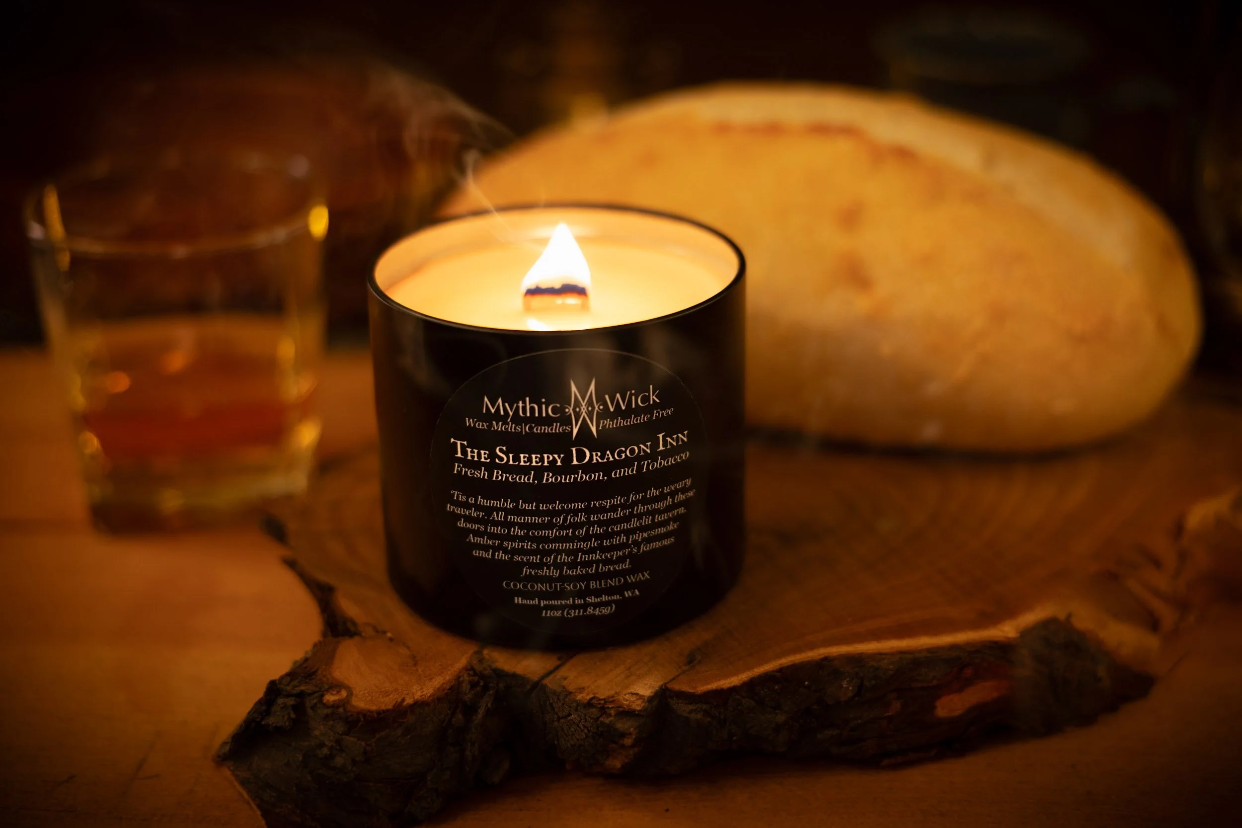 A black candle tin with a wooden wick sits in front of a loaf of rustic bread and a glass of whiskey. 