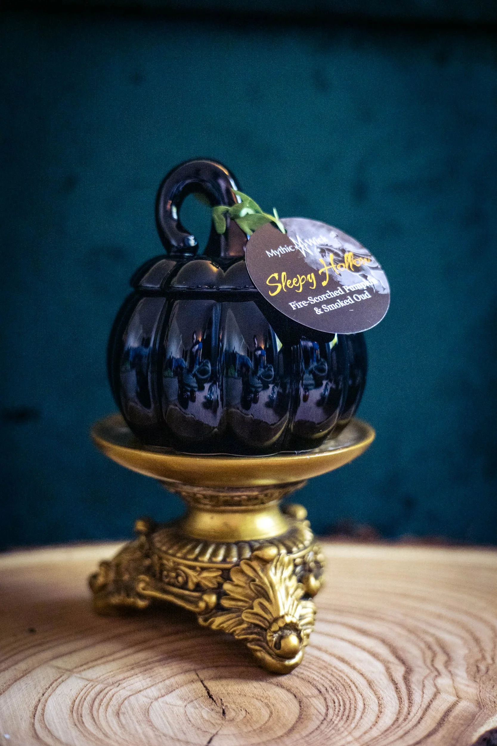 A ceramic black pumpkin jar candle sits on an ornate golden pedestal. The hang tag reads "Sleepy Hollow"