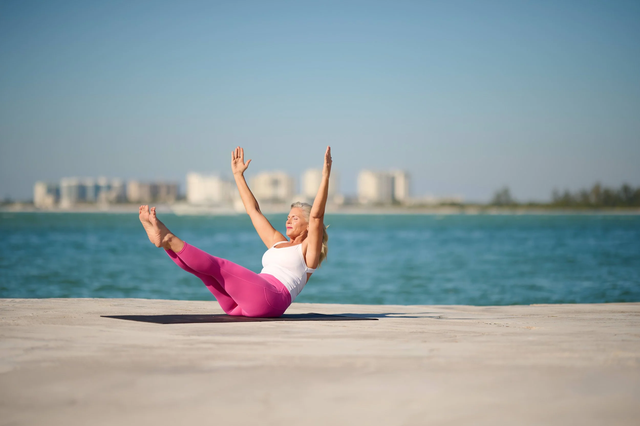 A woman practicing yoga on the beach during the day, sitting on the sand with her legs extended and arms raised, with an ocean beachfront and buildings in the background.