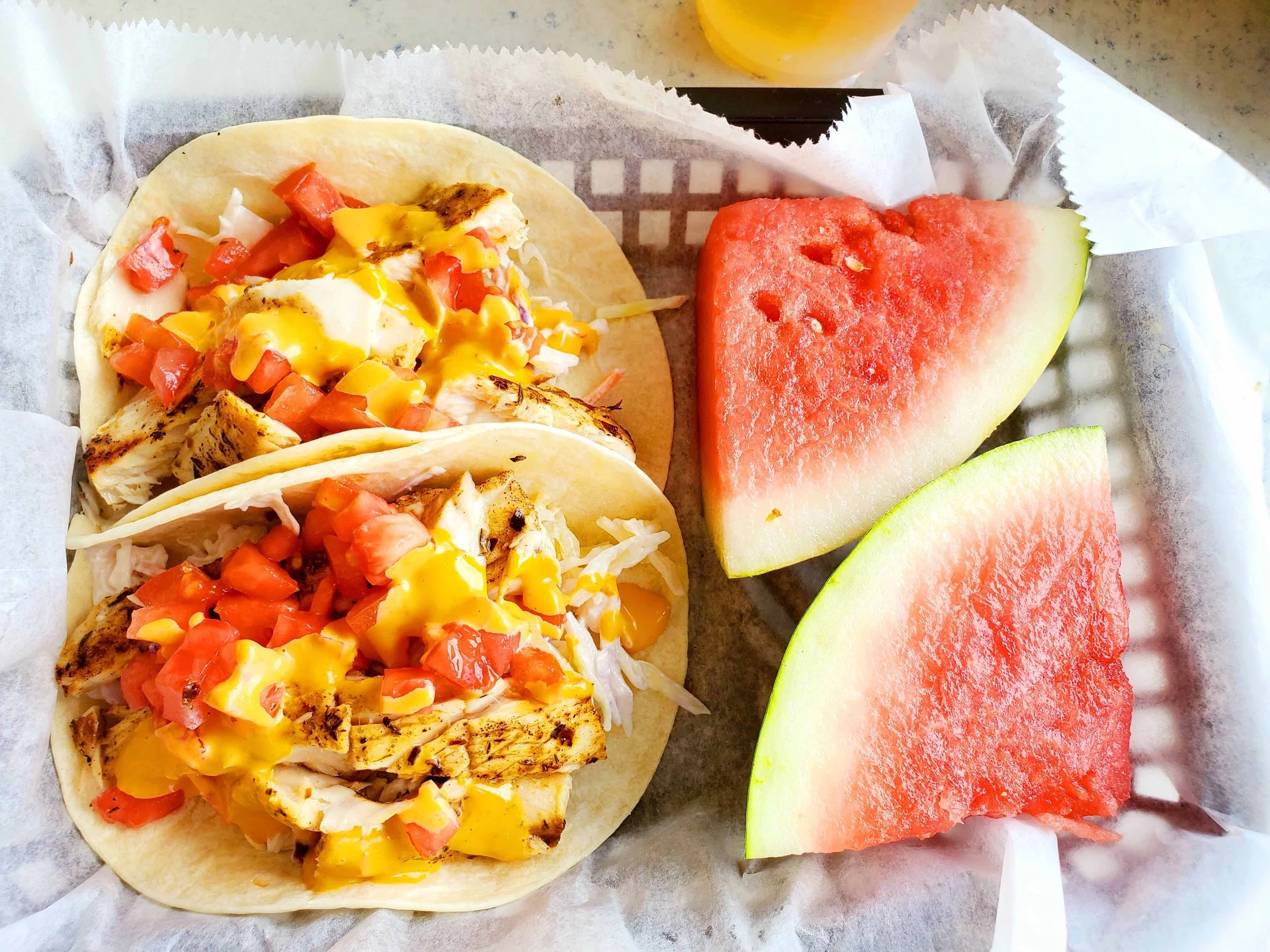 two tacos with chicken, coleslaw, tomatoes, and a yellow sauce, places next to two watermelon slices